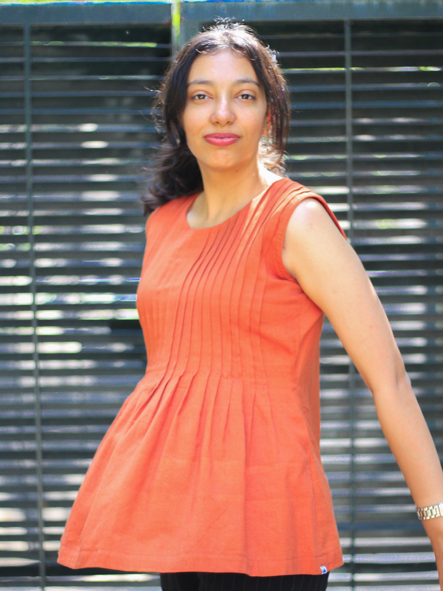 Model wearing Evara’s Rust Reverie Sleeveless Pleated Top, crafted from a sustainable blend of banana fiber and cotton. This airy light blue top features a sleeveless design, fine vertical pleats on the bodice, and a softly flared hem that offers a relaxed yet refined silhouette. Perfect for warm-weather layering or standalone elegance, it blends comfort with conscious fashion. Designed for consumers who value eco-friendly fabrics and contemporary, timeless style. Shop clothing at Evara.