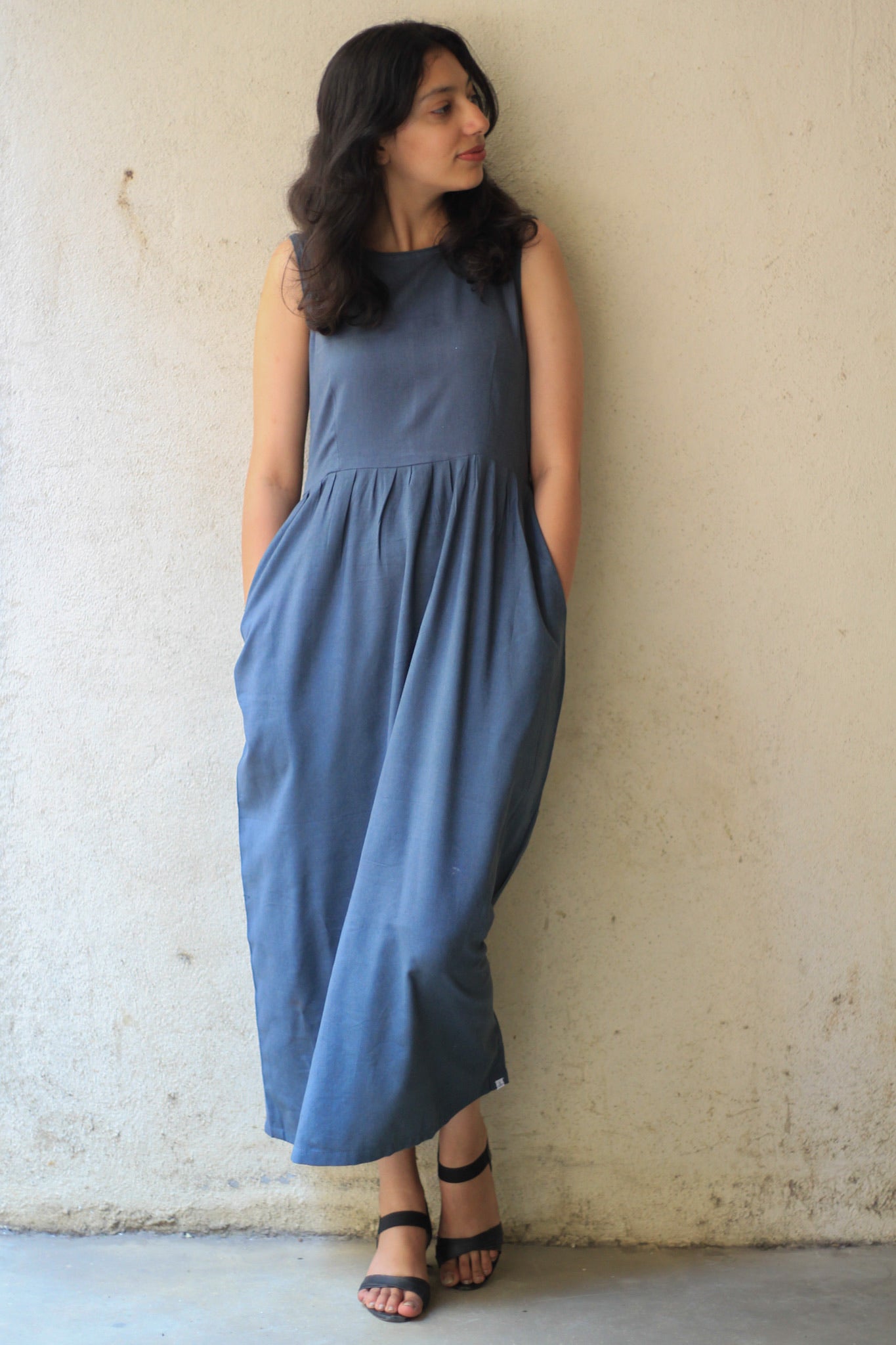 Model wearing Evara’s Cosmic Dusk Sleeveless Dress, crafted from a premium blend of banana fiber and cotton. This elegant midi dress in a deep dusk blue hue features a sleeveless design, a flattering gathered waist, and functional side pockets for convenience. A subtle back slit adds ease of movement, making it a perfect blend of style and comfort. Designed for consumers who appreciate natural fabrics and timeless fashion. Shop clothing at Evara.