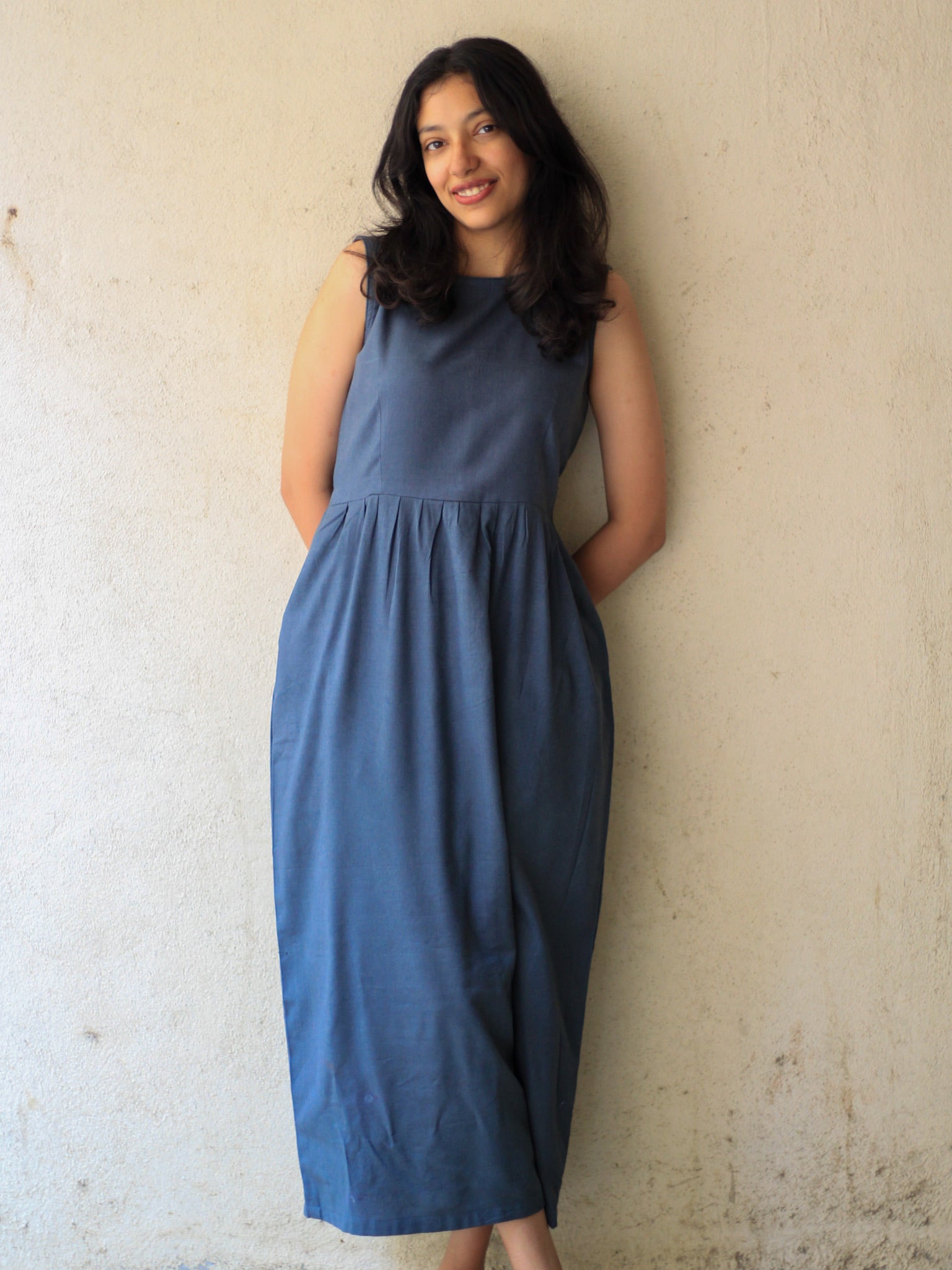 Model wearing Evara’s Cosmic Dusk Sleeveless Dress, crafted from a premium blend of banana fiber and cotton. This elegant midi dress in a deep dusk blue hue features a sleeveless design, a flattering gathered waist, and functional side pockets for convenience. A subtle back slit adds ease of movement, making it a perfect blend of style and comfort. Designed for consumers who appreciate natural fabrics and timeless fashion. Shop clothing at Evara.