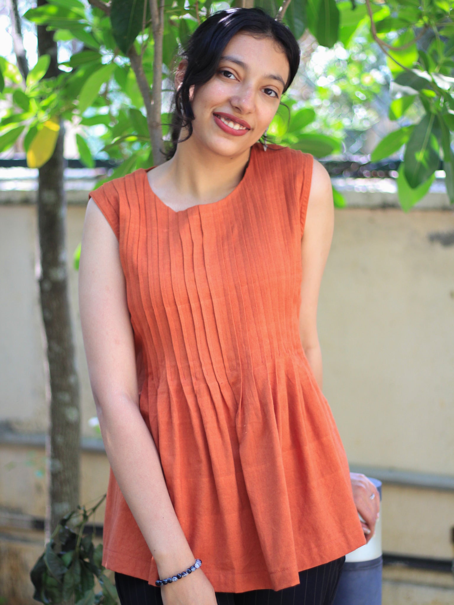 Model wearing Evara’s Rust Reverie Sleeveless Pleated Top, crafted from a sustainable blend of banana fiber and cotton. This airy light blue top features a sleeveless design, fine vertical pleats on the bodice, and a softly flared hem that offers a relaxed yet refined silhouette. Perfect for warm-weather layering or standalone elegance, it blends comfort with conscious fashion. Designed for consumers who value eco-friendly fabrics and contemporary, timeless style. Shop clothing at Evara.