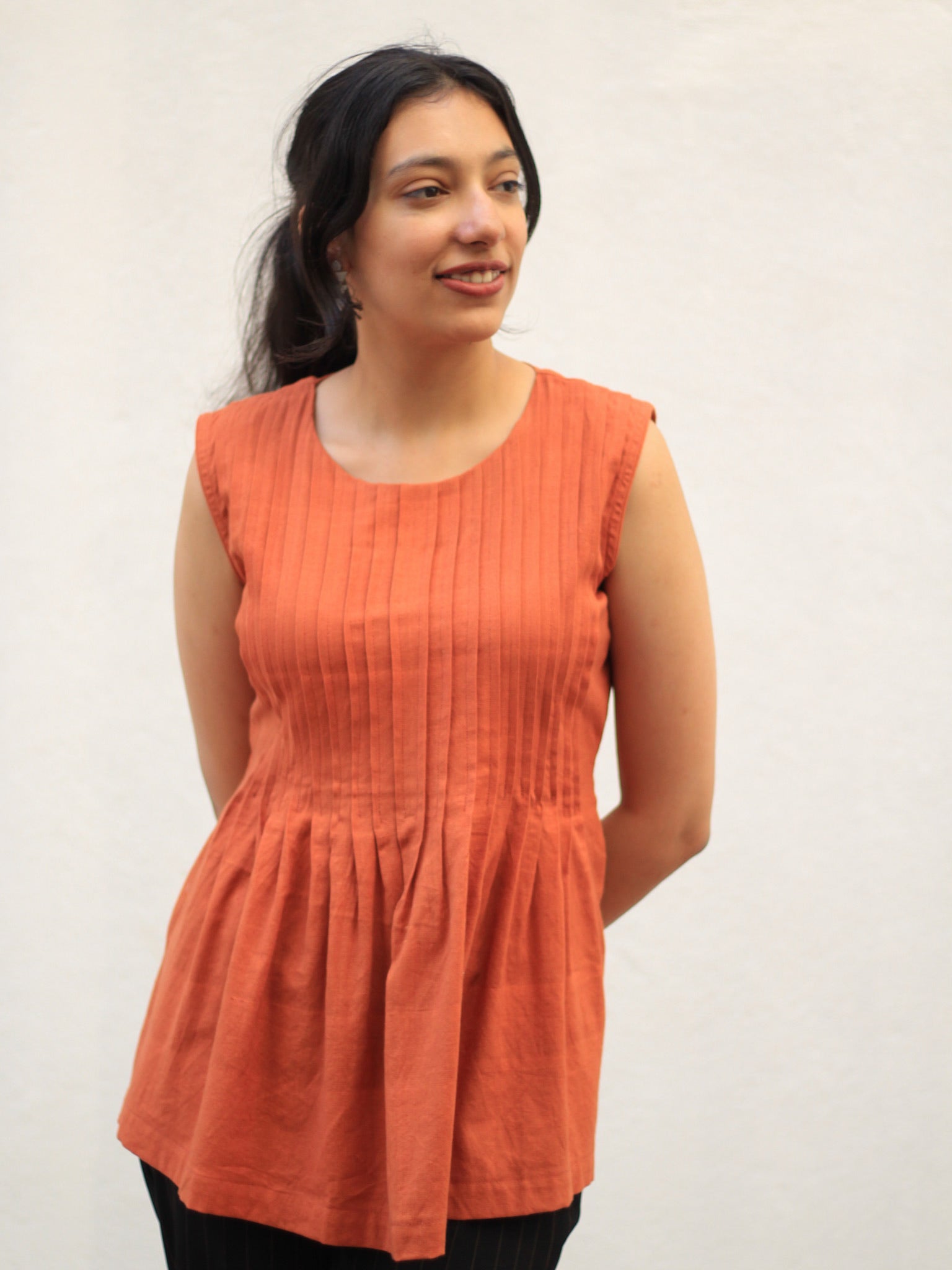 Model wearing Evara’s Rust Reverie Sleeveless Pleated Top, crafted from a sustainable blend of banana fiber and cotton. This airy light blue top features a sleeveless design, fine vertical pleats on the bodice, and a softly flared hem that offers a relaxed yet refined silhouette. Perfect for warm-weather layering or standalone elegance, it blends comfort with conscious fashion. Designed for consumers who value eco-friendly fabrics and contemporary, timeless style. Shop clothing at Evara.