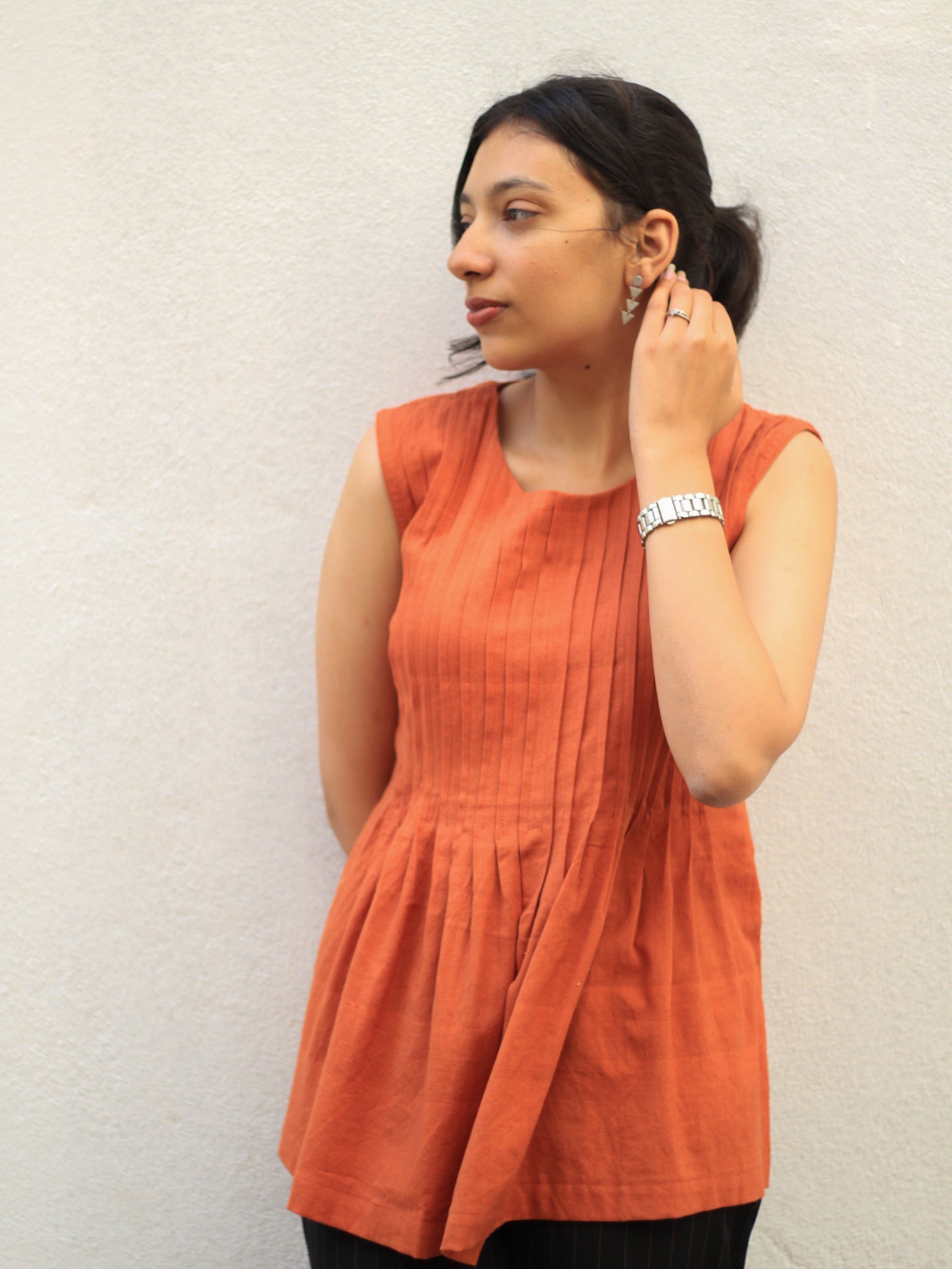 Model wearing Evara’s Rust Reverie Sleeveless Pleated Top, crafted from a sustainable blend of banana fiber and cotton. This airy light blue top features a sleeveless design, fine vertical pleats on the bodice, and a softly flared hem that offers a relaxed yet refined silhouette. Perfect for warm-weather layering or standalone elegance, it blends comfort with conscious fashion. Designed for consumers who value eco-friendly fabrics and contemporary, timeless style. Shop clothing at Evara.