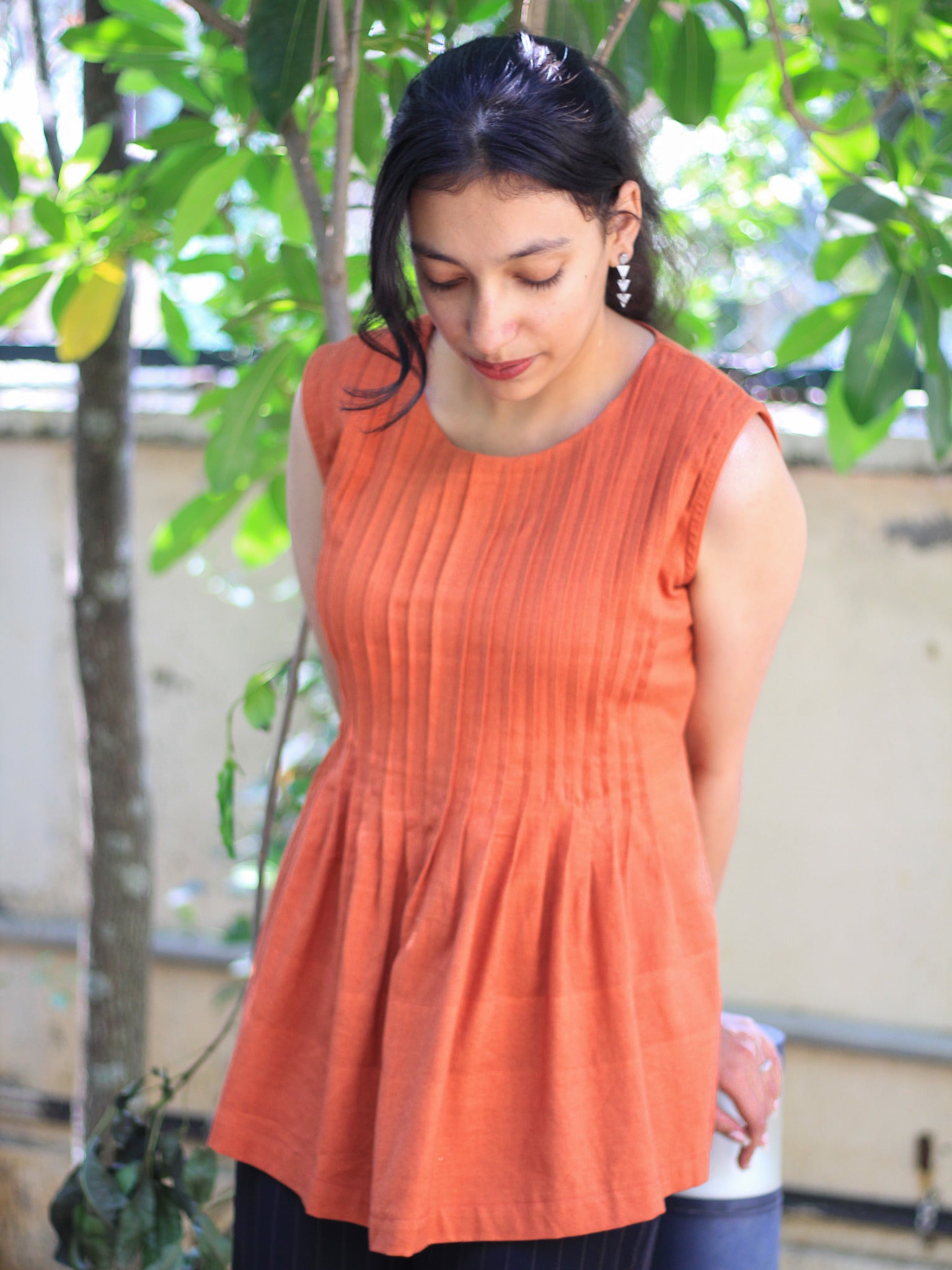 Model wearing Evara’s Rust Reverie Sleeveless Pleated Top, crafted from a sustainable blend of banana fiber and cotton. This airy light blue top features a sleeveless design, fine vertical pleats on the bodice, and a softly flared hem that offers a relaxed yet refined silhouette. Perfect for warm-weather layering or standalone elegance, it blends comfort with conscious fashion. Designed for consumers who value eco-friendly fabrics and contemporary, timeless style. Shop clothing at Evara.