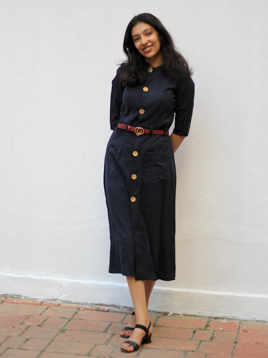 Model wearing Evara’s Obsidian Noir Button-Down Dress, crafted from a premium blend of banana fiber and cotton. This timeless black midi dress features a structured silhouette, three-quarter sleeves, a classic button-down front, and functional patch pockets. Styled with a belt for a defined waist, it offers effortless sophistication for work or casual outings. Designed for consumers who appreciate natural fabrics and timeless fashion. Shop clothing at Evara.