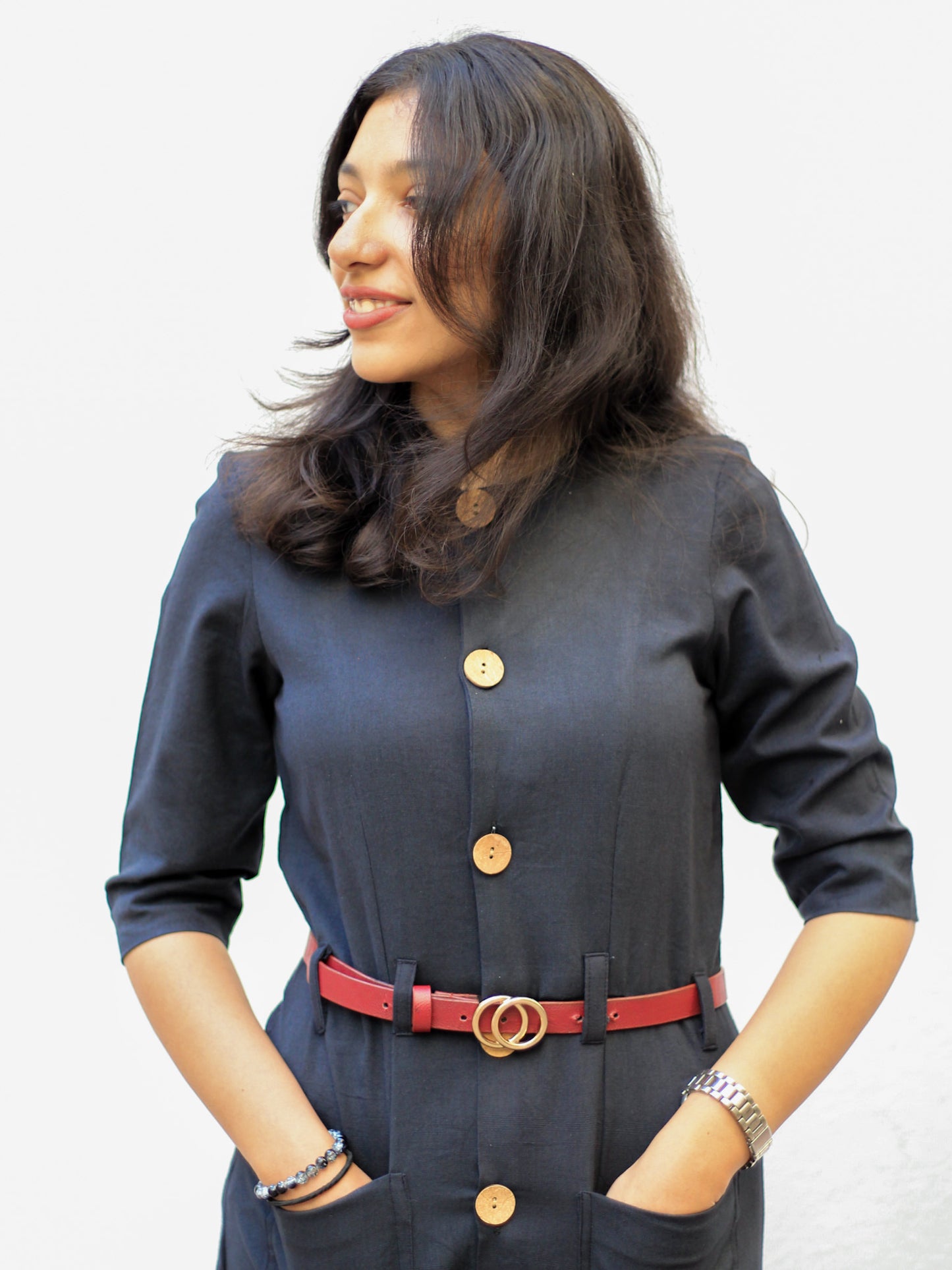 Model wearing Evara’s Obsidian Noir Button-Down Dress, crafted from a premium blend of banana fiber and cotton. This timeless black midi dress features a structured silhouette, three-quarter sleeves, a classic button-down front, and functional patch pockets. Styled with a belt for a defined waist, it offers effortless sophistication for work or casual outings. Designed for consumers who appreciate natural fabrics and timeless fashion. Shop clothing at Evara.
