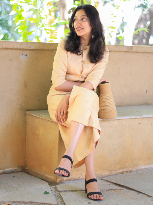 Model wearing Evara’s Golden Aura Button-Down Dress, crafted from a premium blend of banana fiber and cotton. This elegant midi dress in a warm golden hue features a structured silhouette, three-quarter sleeves, a classic button-down front, and functional patch pockets. Styled with a belt for a defined waist, it offers effortless sophistication for work or casual outings. Designed for consumers who appreciate natural fabrics and timeless fashion. Shop clothing at Evara.