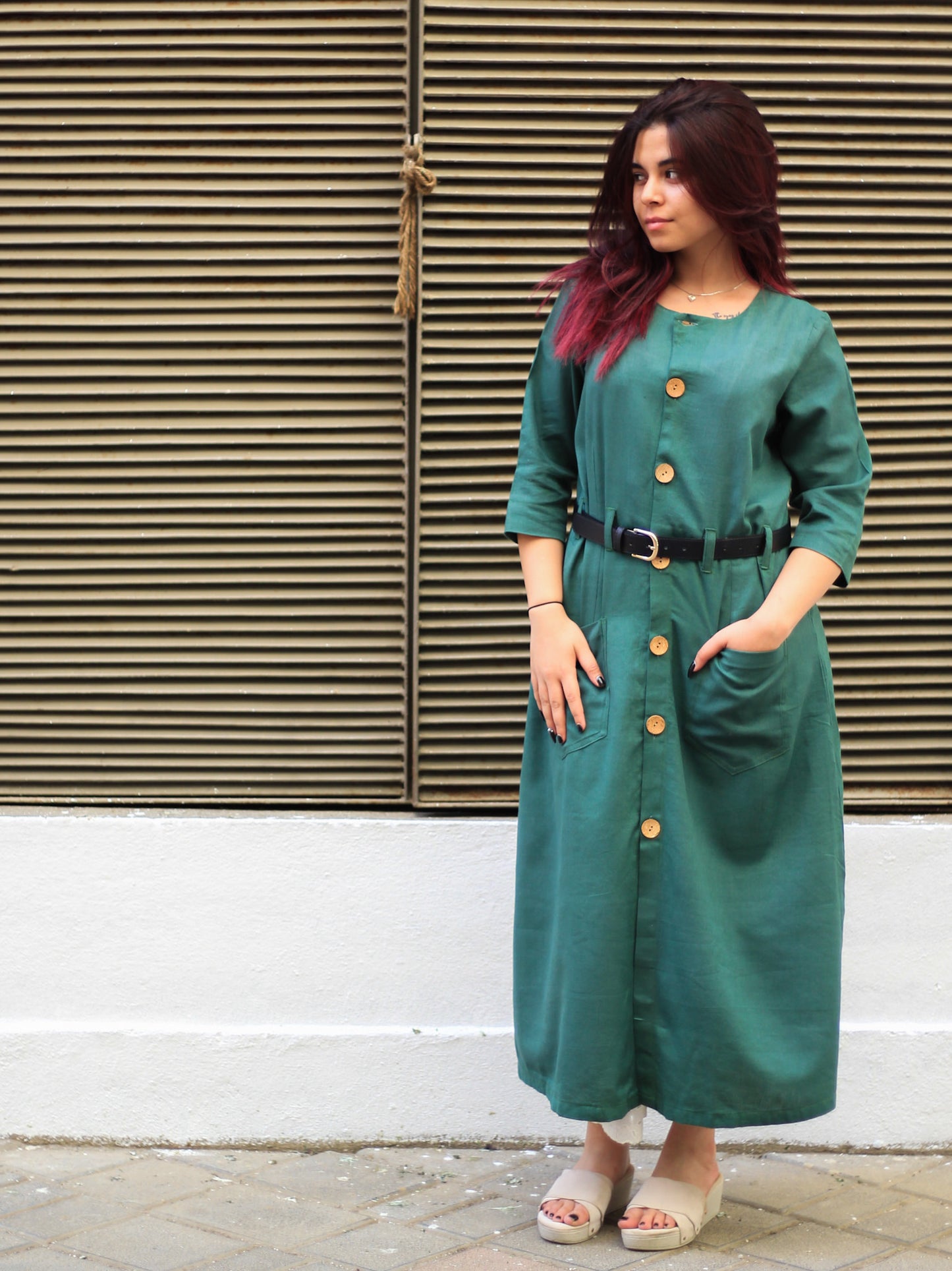 Model wearing Evara’s Botanic Luxe Button-Down Dress, crafted from a premium blend of banana fiber and cotton. This elegant midi dress in a green hue features a structured silhouette, three-quarter sleeves, a classic button-down front, and functional patch pockets. Styled with a belt for a defined waist, it offers effortless sophistication for work or casual outings. Designed for consumers who appreciate natural fabrics and timeless fashion. Shop clothing at Evara.