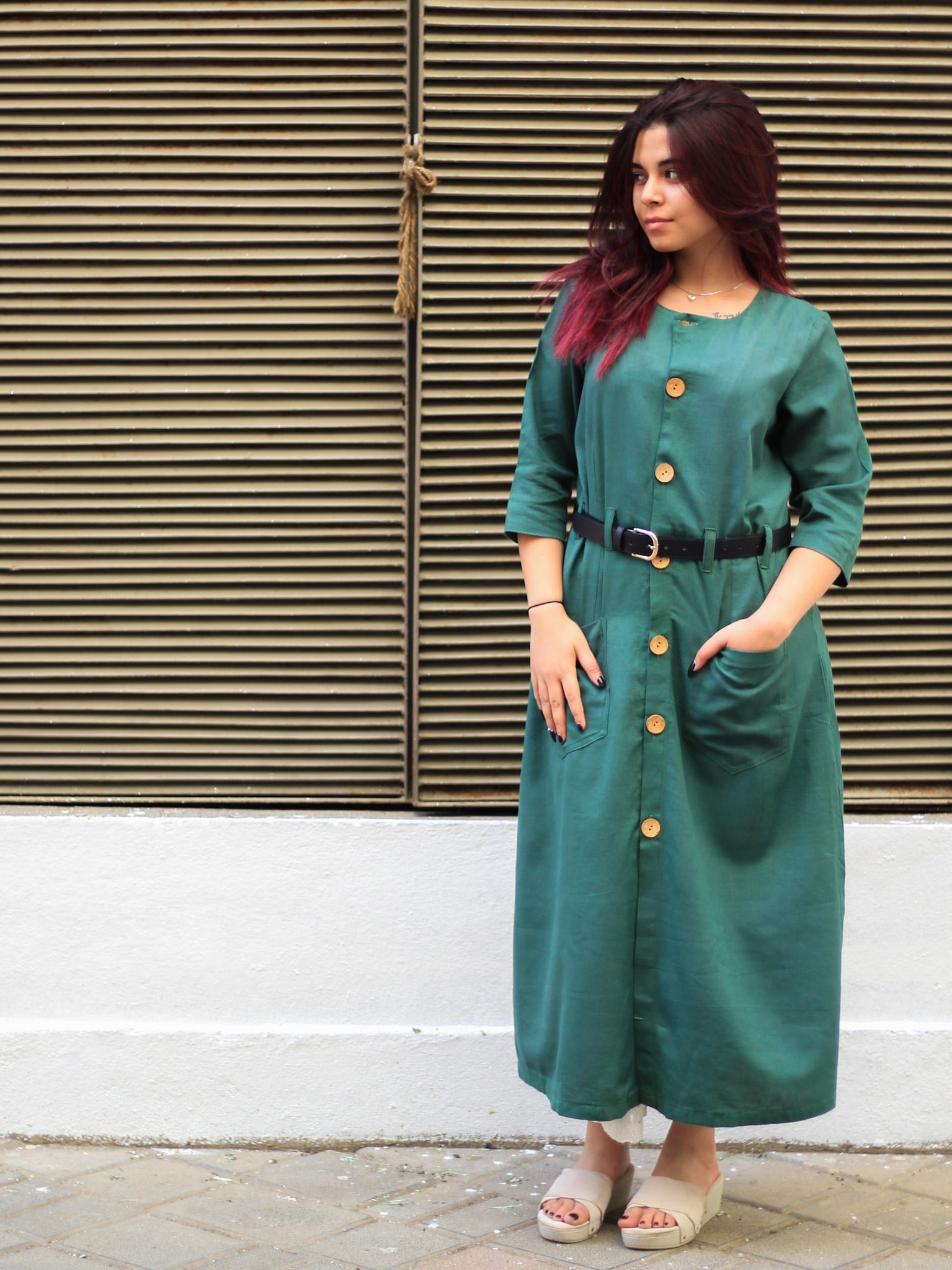 Model wearing Evara’s Botanic Luxe Button-Down Dress, crafted from a premium blend of banana fiber and cotton. This elegant midi dress in a green hue features a structured silhouette, three-quarter sleeves, a classic button-down front, and functional patch pockets. Styled with a belt for a defined waist, it offers effortless sophistication for work or casual outings. Designed for consumers who appreciate natural fabrics and timeless fashion. Shop clothing at Evara.