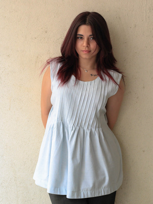 Model wearing Evara’s Skyline Haze Sleeveless Pleated Top, crafted from a sustainable blend of banana fiber and cotton. This airy light blue top features a sleeveless design, fine vertical pleats on the bodice, and a softly flared hem that offers a relaxed yet refined silhouette. Perfect for warm-weather layering or standalone elegance, it blends comfort with conscious fashion. Designed for consumers who value eco-friendly fabrics and contemporary, timeless style. Shop clothing at Evara.